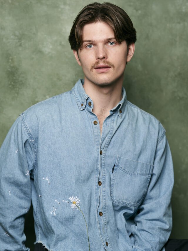A young man with brown hair, a mustache, and blue eyes wearing a light blue denim shirt against a green background.