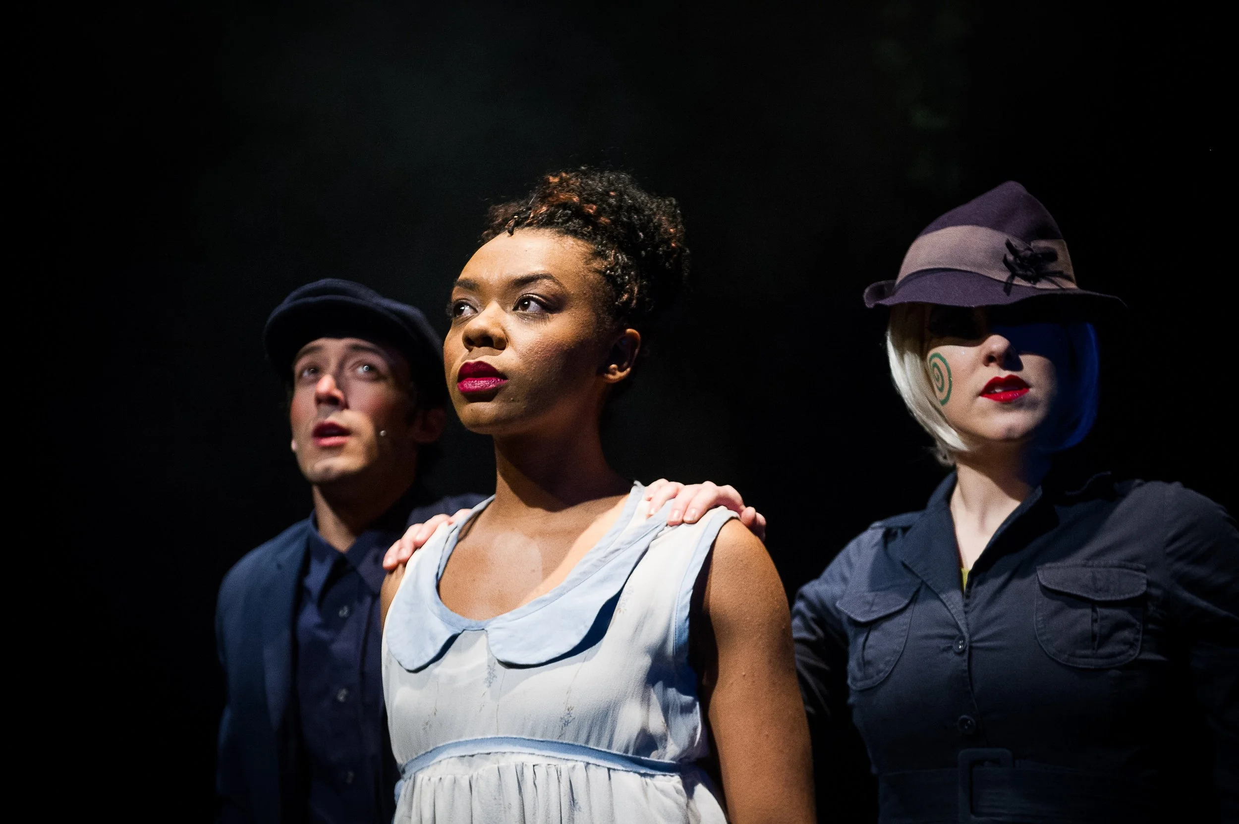Three actors on stage in a theatrical performance, with two women and one man, standing close together against a dark background. The woman in the center has dark skin, curly hair styled up, and wears a white dress with a rounded collar. The woman on the right has light skin, platinum blonde hair, wears a wide-brimmed hat, dark shirt, and has spiraled face paint. The man on the left has light skin, wears a dark hat and dark shirt, with a serious expression.