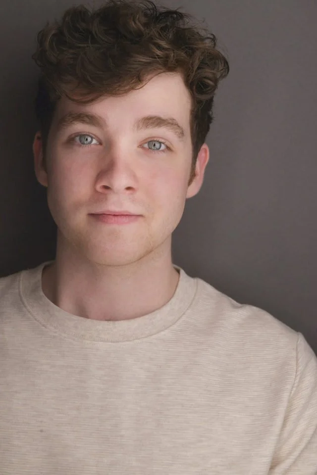 Close-up portrait of a young man with curly brown hair, blue eyes, and fair skin, wearing a beige crewneck shirt, against a gray background.
