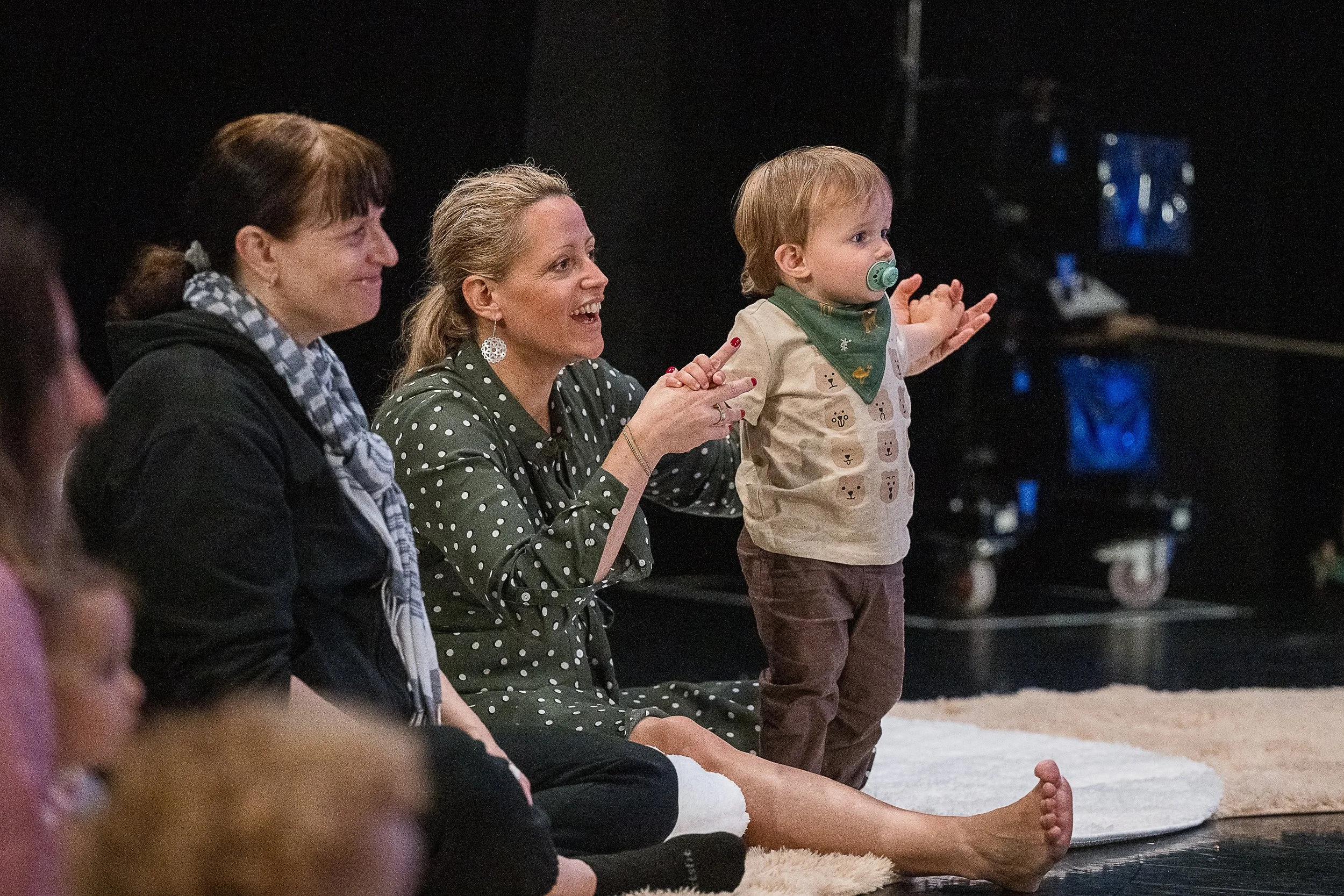 Adults and a young child sitting on a carpeted floor, watching and clapping, with a dark background and a television screen in the distance.