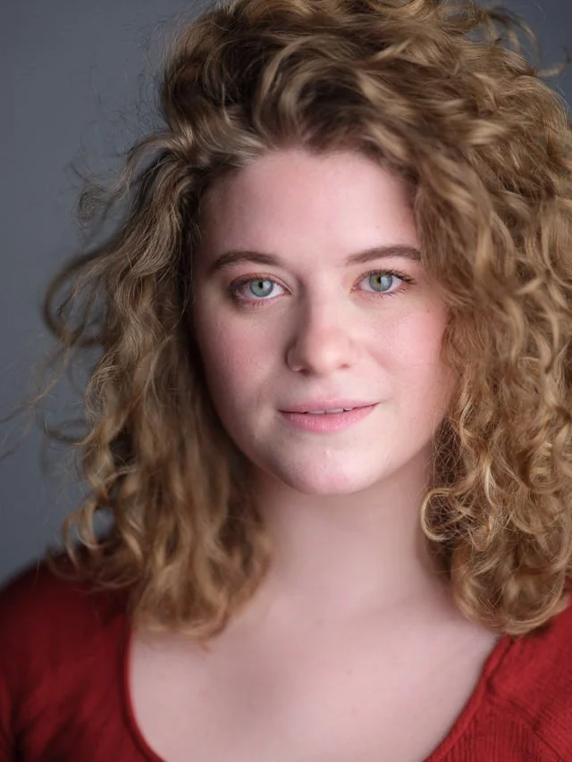 Close-up portrait of a young woman with curly red hair and blue eyes, wearing a red top, against a plain neutral background.