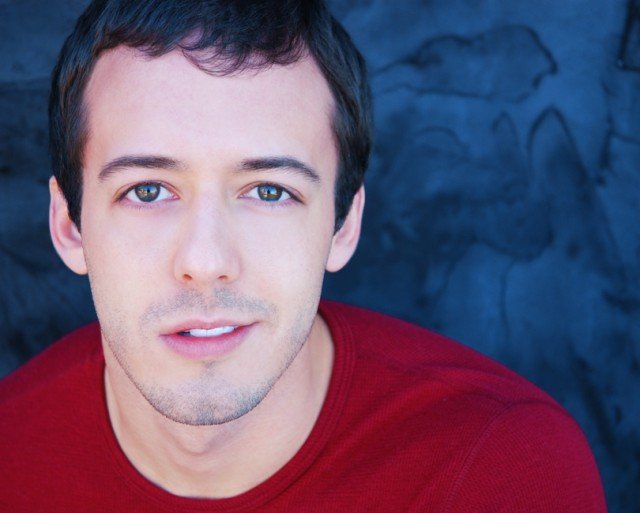 Close-up portrait of a young man with short dark hair, blue eyes, and light skin, wearing a red shirt, with a dark, textured background.