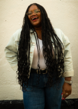 Woman with long dreadlocks, glasses, wearing a white jacket and jeans, laughing against a white wall.