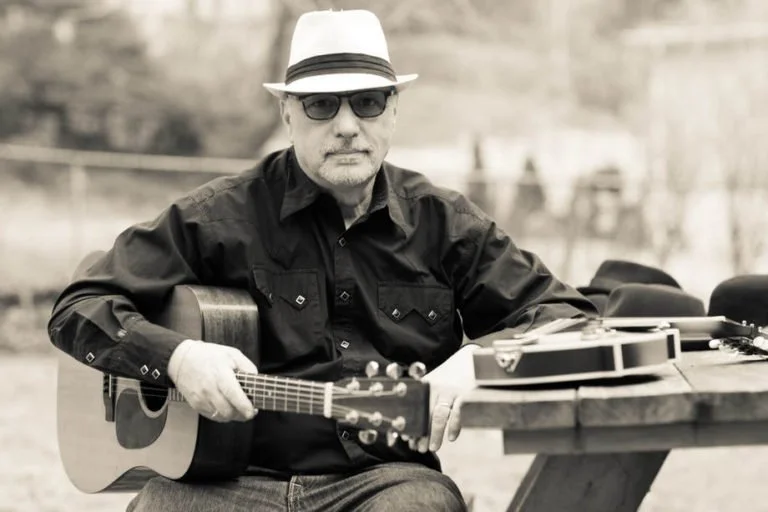 Man wearing a fedora hat, sunglasses, and a dark shirt, sitting outdoors and holding an acoustic guitar.