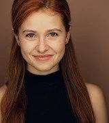 A young woman with red hair smiling, wearing a black top against a neutral background.