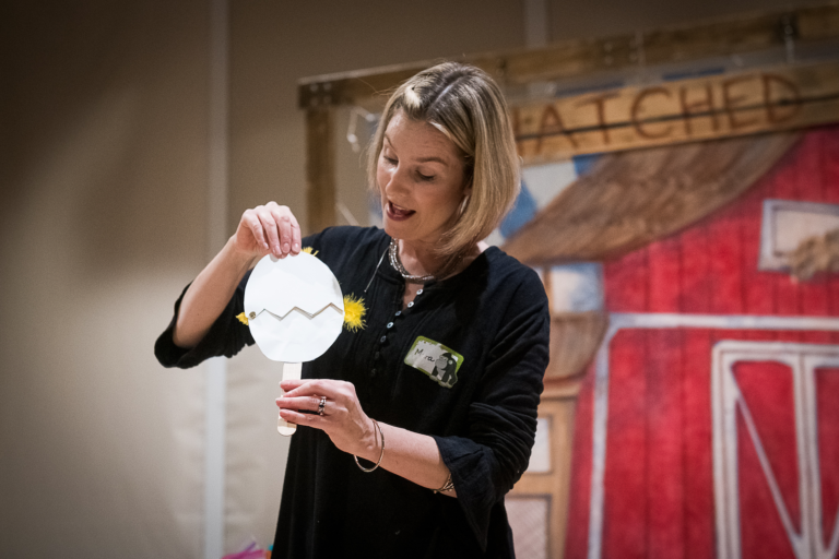 Woman holding a paper craft that looks like a cracked egg, standing in front of a barn-themed background.