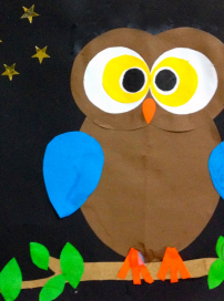 A colorful paper craft of an owl with big eyes, blue wings, orange beak, and green leaves at the bottom, set against a black background with yellow stars.