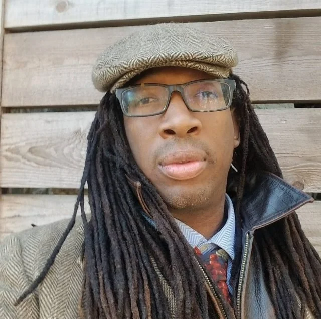 A man with long dreadlocks, wearing glasses, a beige flat cap, a patterned tie, and a leather jacket, sitting in front of a wooden wall.