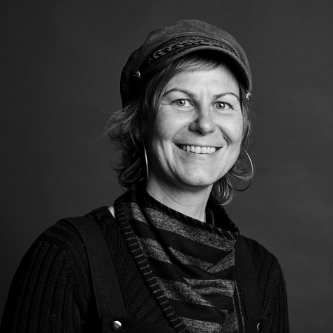 Black and white portrait of a smiling woman wearing a cap, striped scarf, and dark clothing.