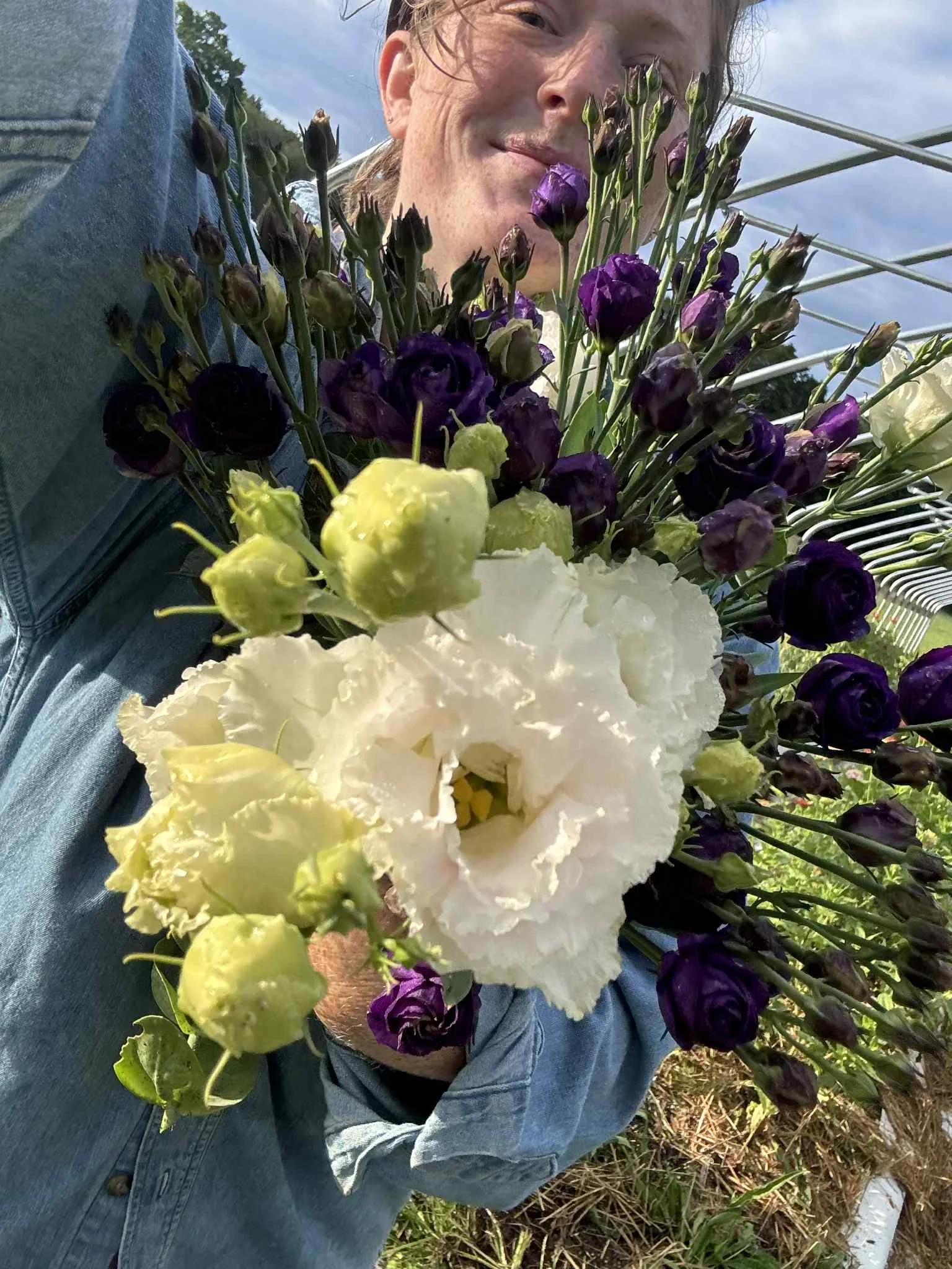 Person holding a large bouquet of white and purple flowers outdoors, with a partly cloudy sky in the background.