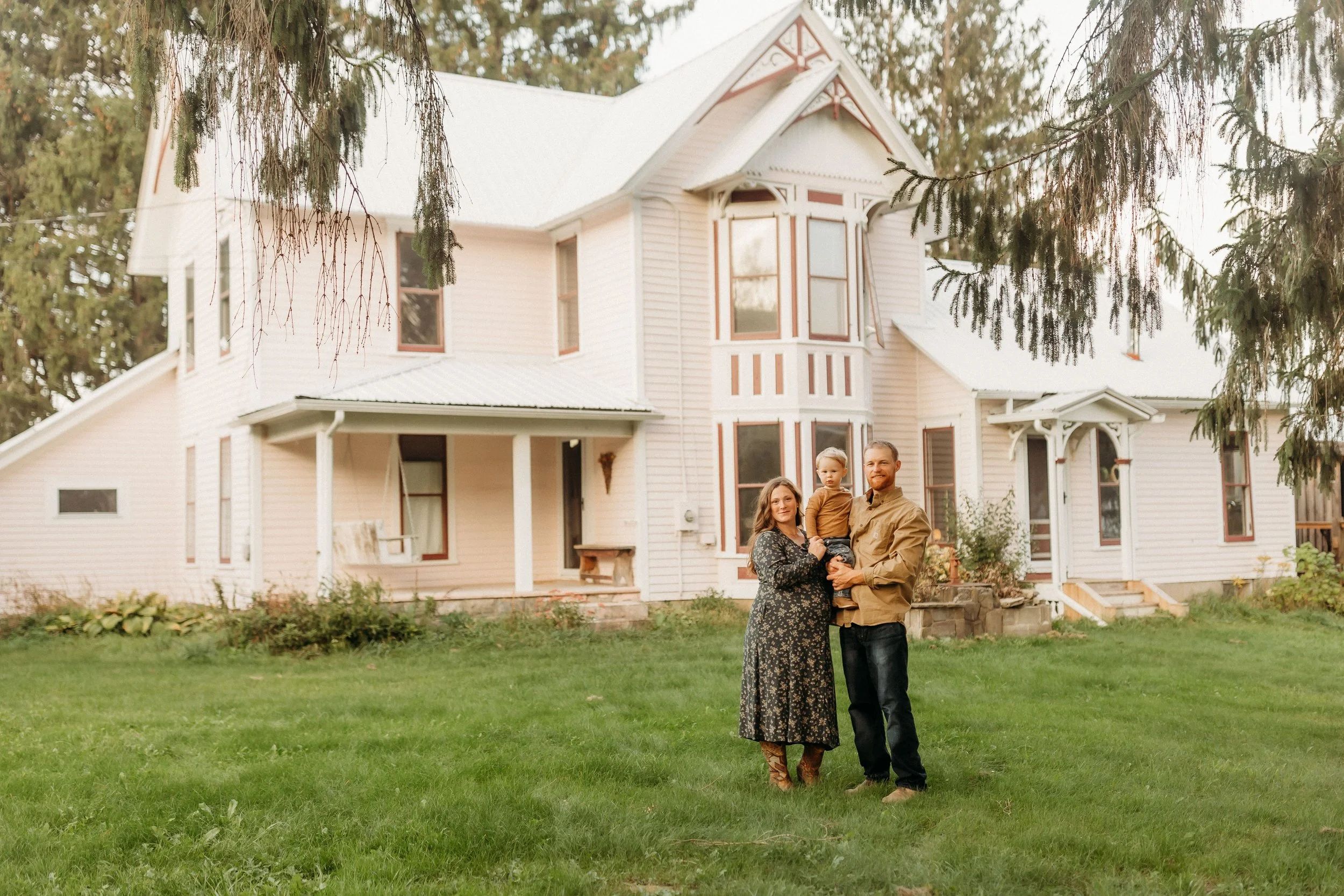 A family of three standing on a lawn in front of a large, white, Victorian-style house with multiple windows and a porch, surrounded by trees.