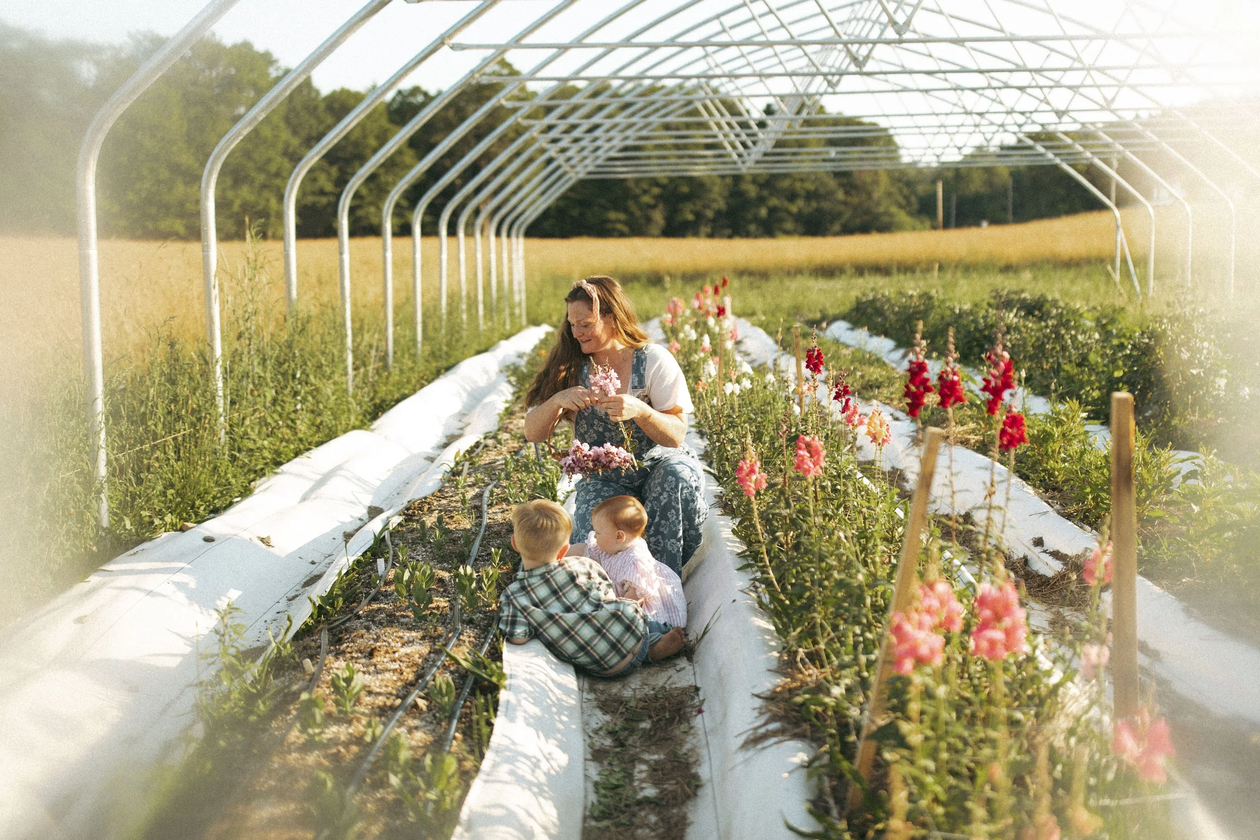 A woman with two young children sitting in a flower garden under a white metal greenhouse frame, surrounded by blooming pink, red, and white flowers, during sunny weather, with green fields and trees in the background.