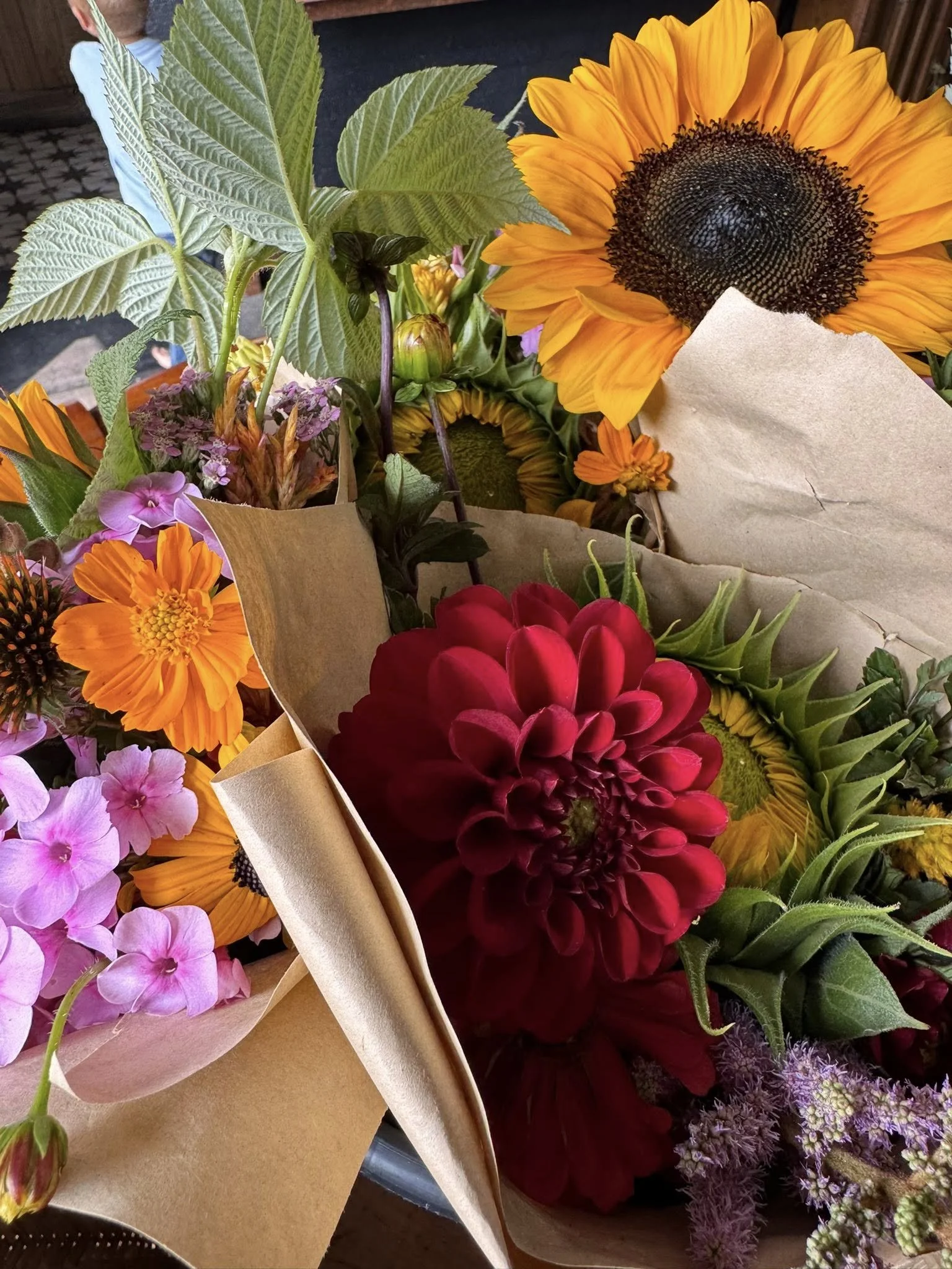 A bouquet of assorted flowers including sunflowers, a large red dahlia, orange zinnias, pink petunias, and purple flowers wrapped in brown paper.