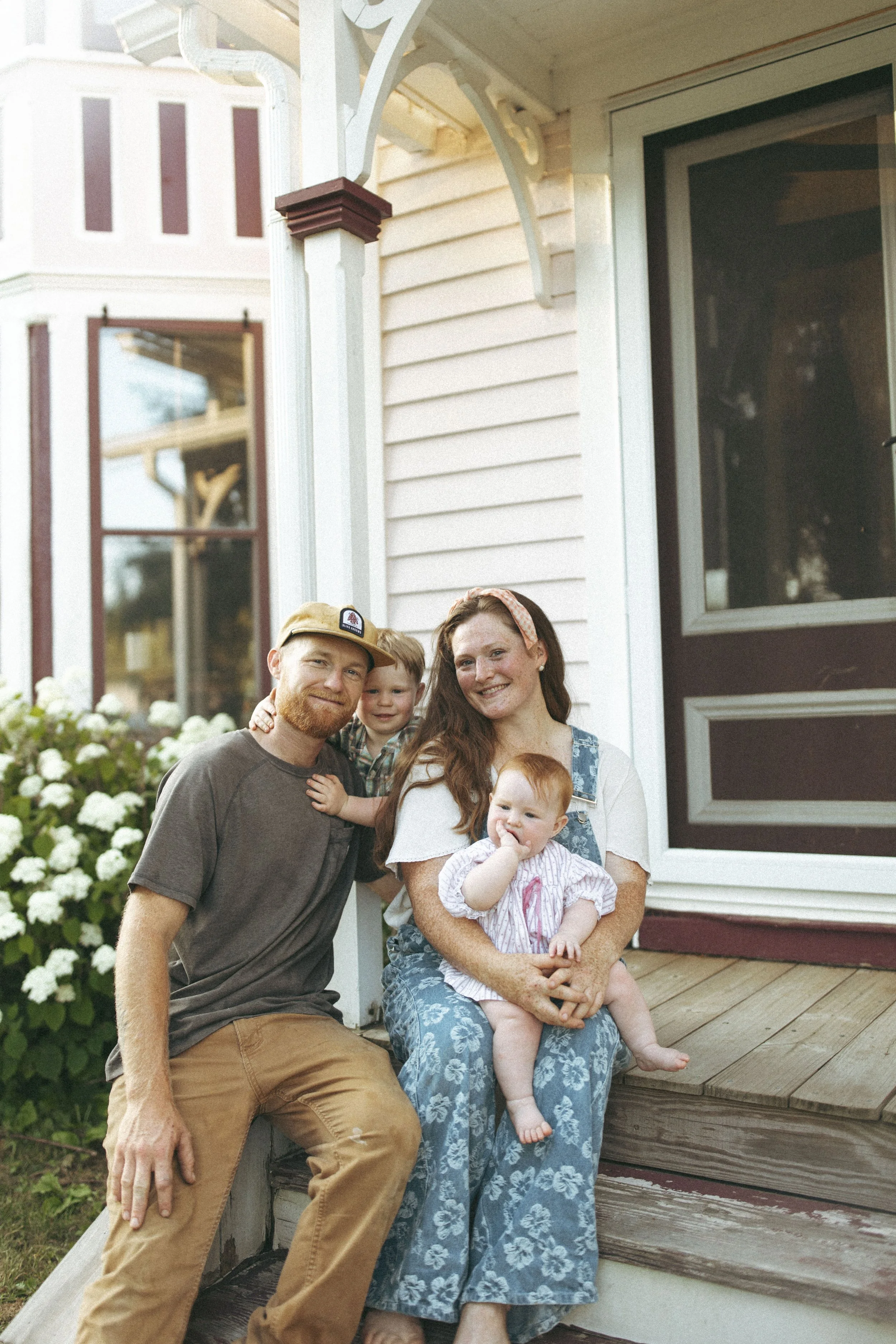 A family of four sitting on the front porch of a house. The mother is holding a baby girl, and a father and young boy are beside them. The house has a wooden deck, white siding, and dark-framed windows.