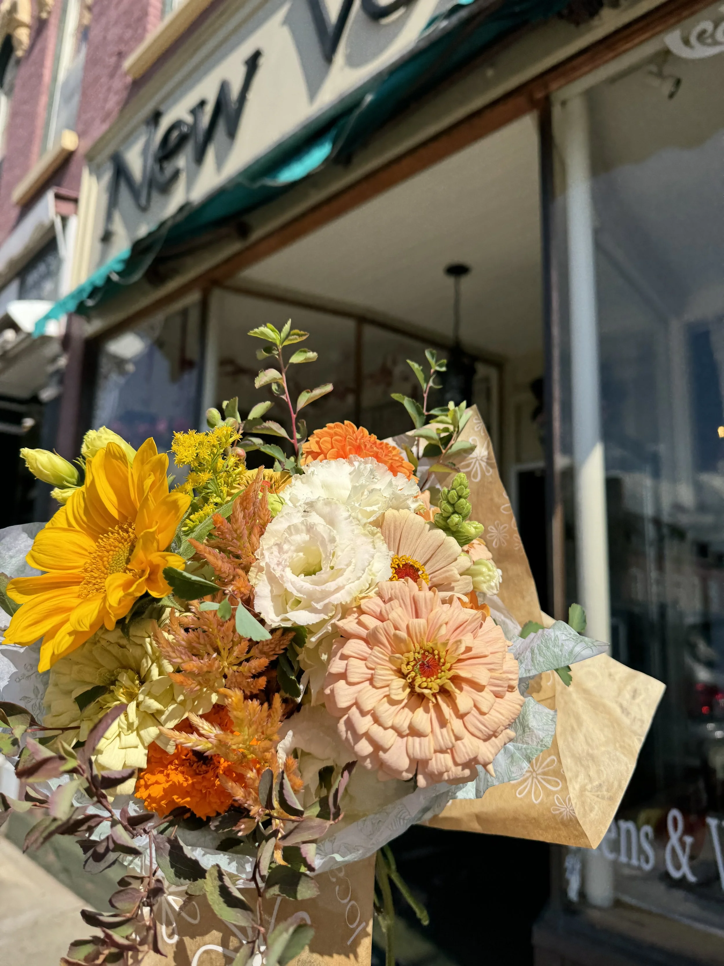 A colorful bouquet of flowers including orange, yellow, and white blooms, held outside a shop with a sign overhead.