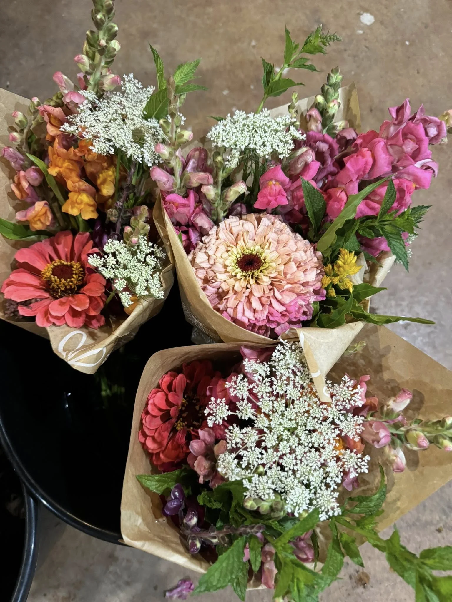 Three bouquets of mixed colorful flowers, including pink, orange, and white blooms with green leaves, wrapped in brown paper.