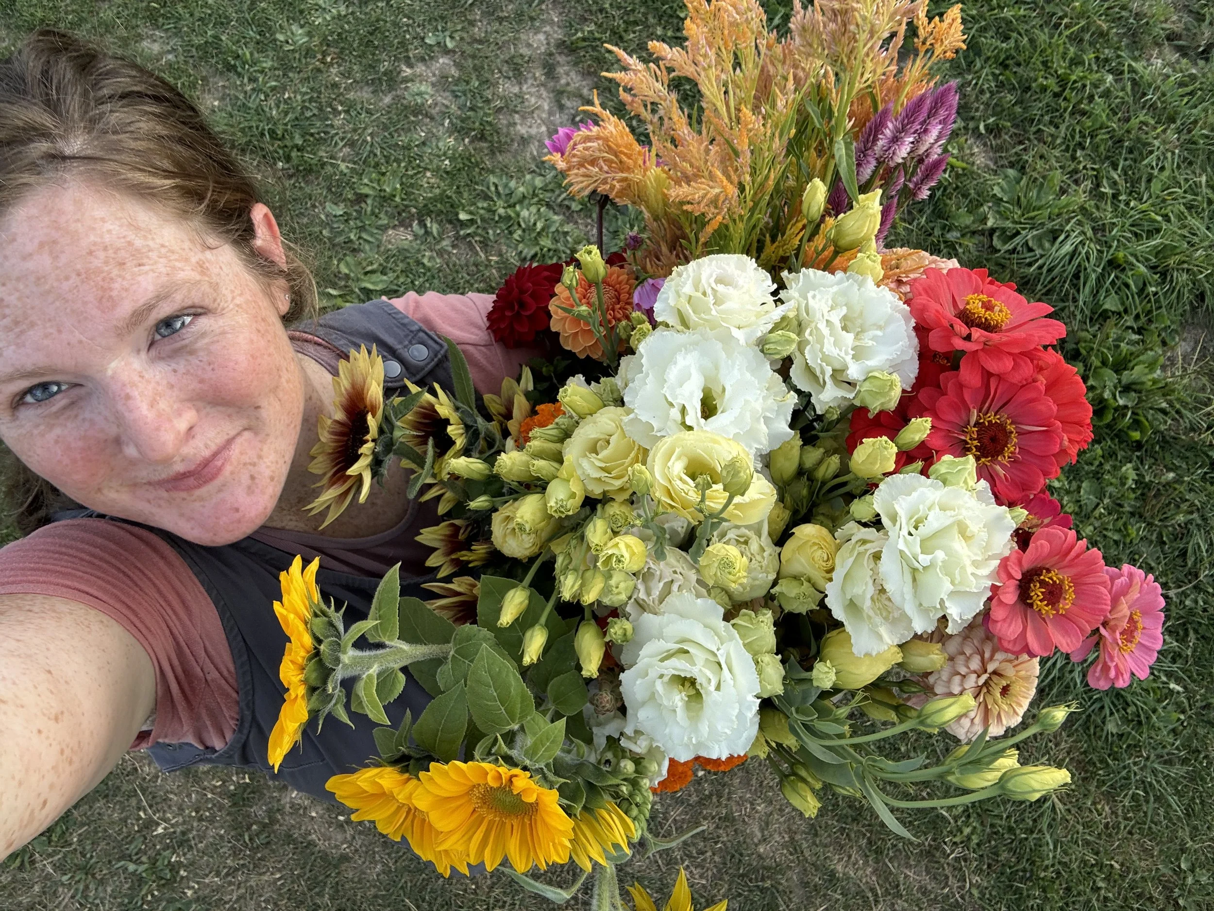 A woman with freckles and light blue eyes taking a selfie outdoors, surrounded by a large bouquet of colorful flowers including white, yellow, red, pink, orange, and purple blossoms, with green grass in the background.