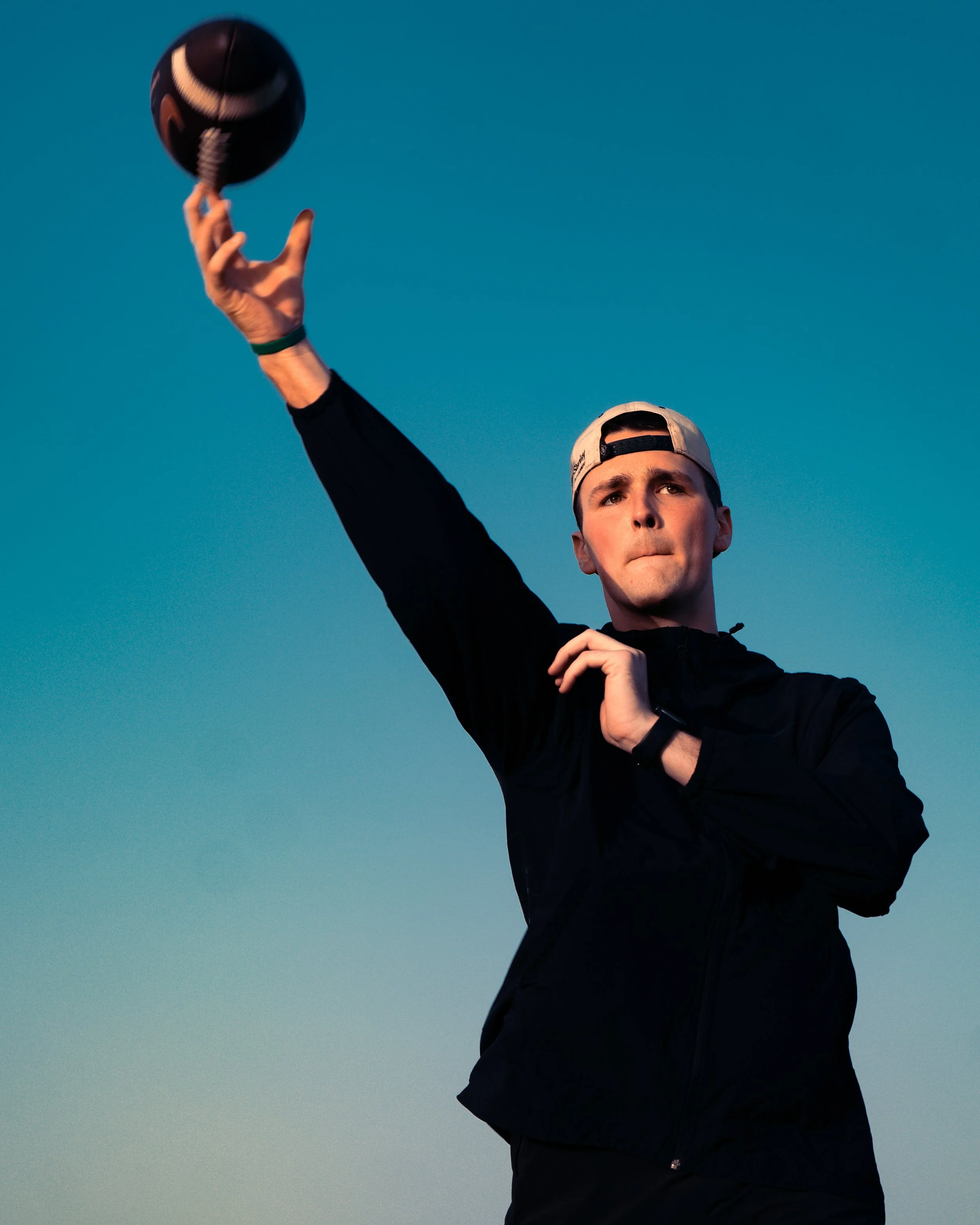 Trey Owens in black jacket and backwards cap throwing a black and white football during sunset with a clear blue sky background.
