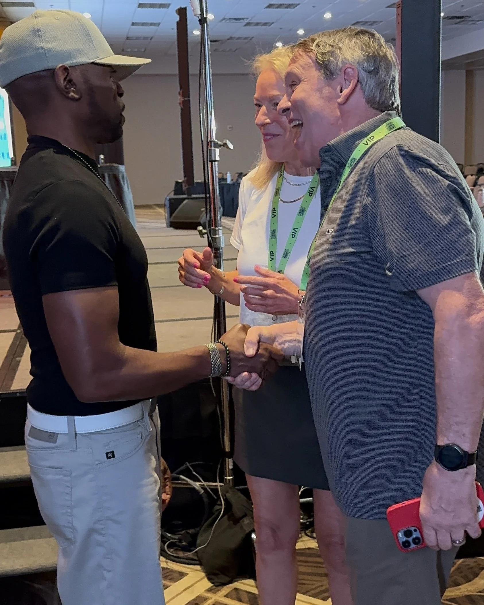 Three individuals engaged in conversation at an event, with two men shaking hands and a woman standing between them, all wearing VIP lanyards.