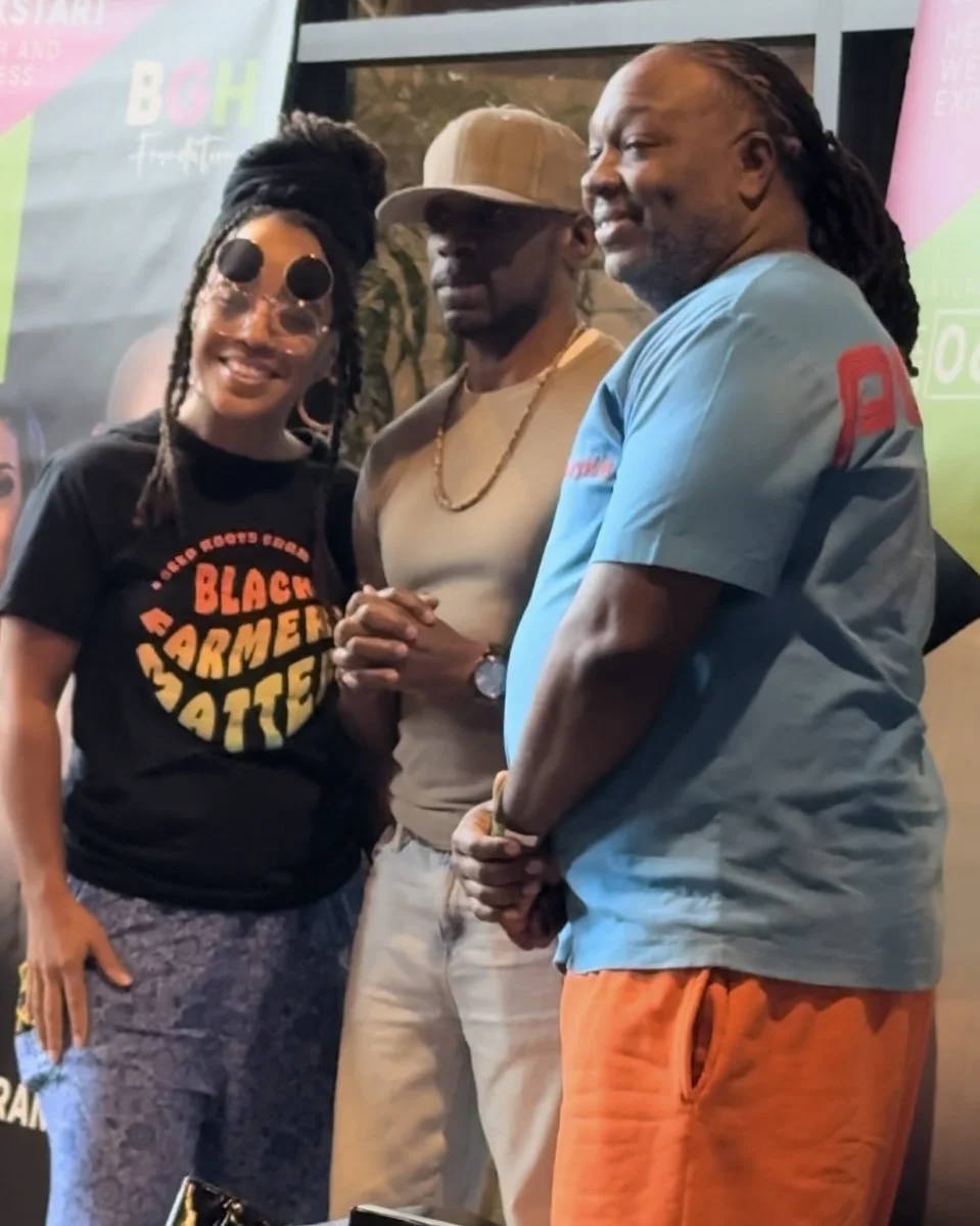 Three people are standing together, smiling. The woman on the left wears sunglasses and a black t-shirt with the words 'Black Farmers Matter'. The man in the middle wears a tan cap and beige clothing, holding hands with the woman. The man on the righ