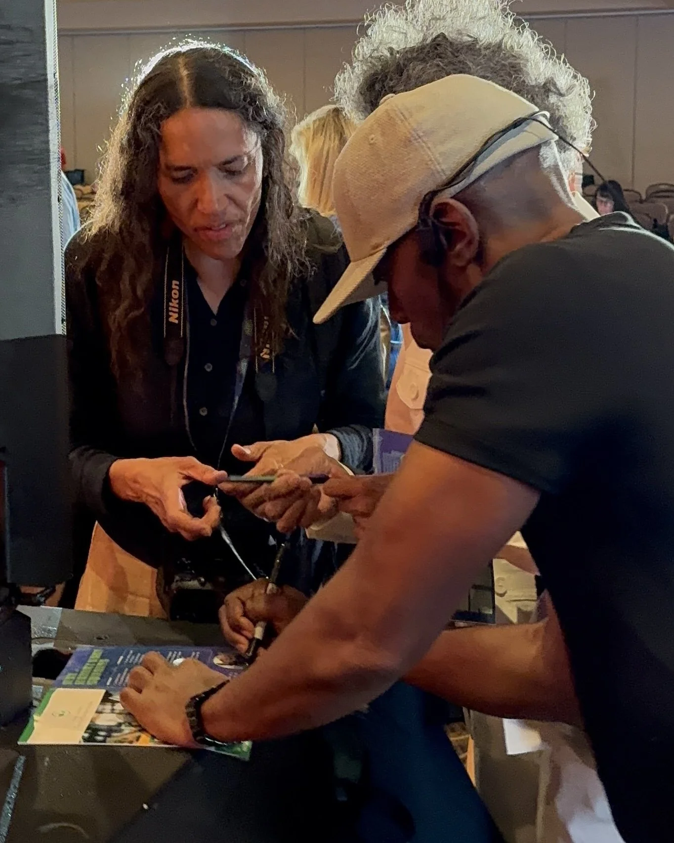 Two people are closely examining a smartphone at a table, with a camera hanging from the woman's neck. There are brochures and papers on the table, and other people in the background.