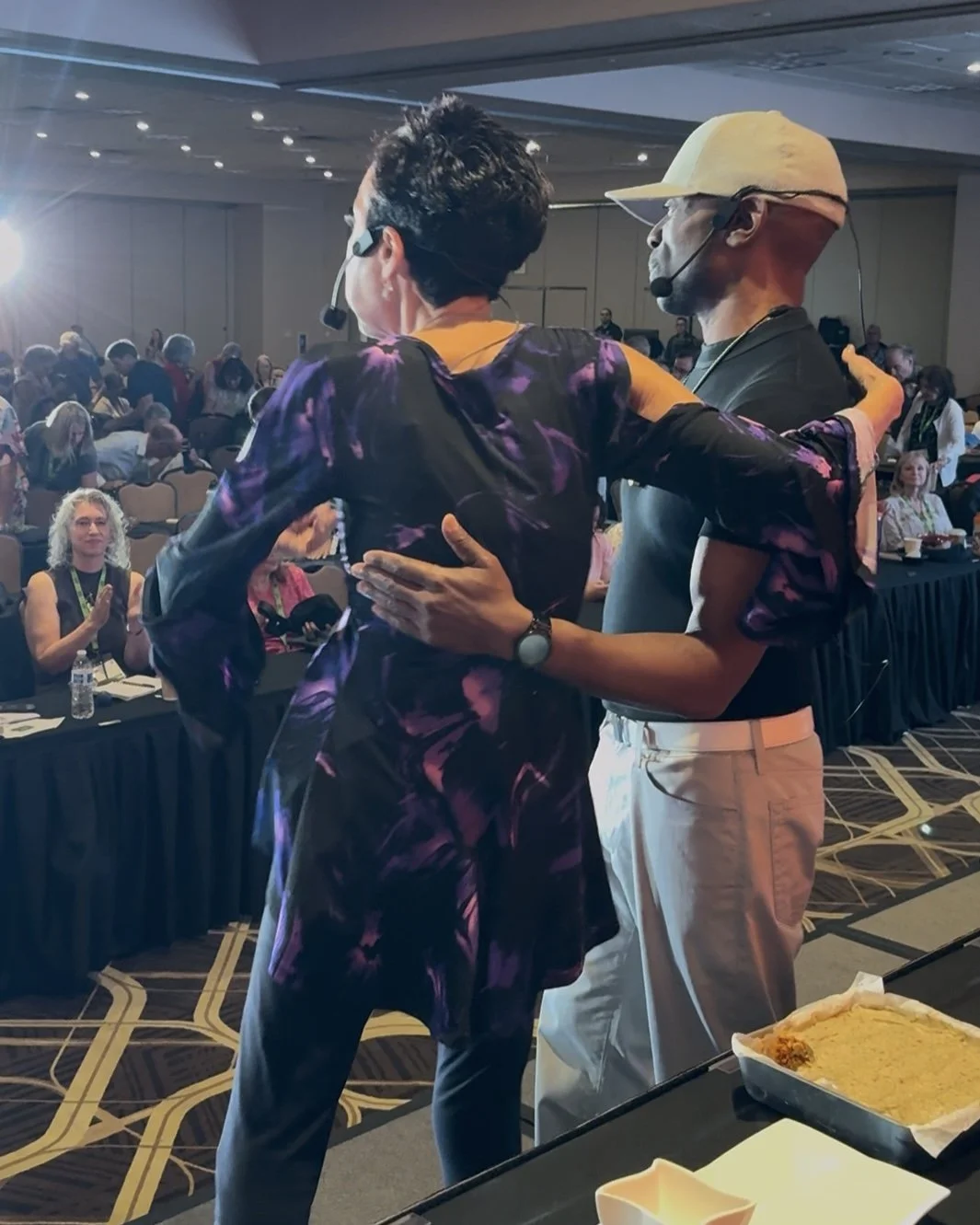 Two speakers on stage hug each other while presenting at a conference, with an audience seated at tables watching.