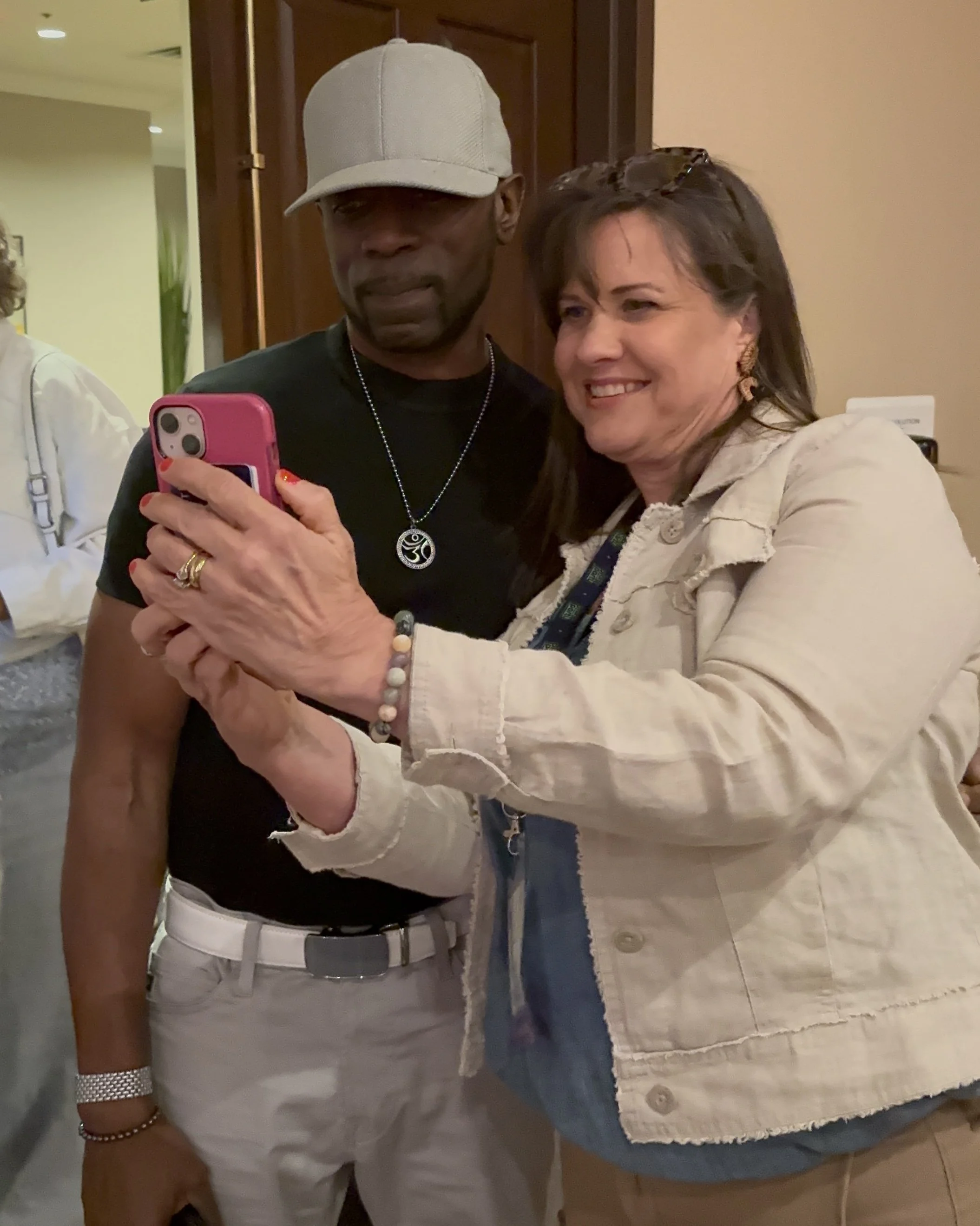 A woman and a man taking a selfie together in an indoor setting. The woman is smiling and holding a pink smartphone, while the man is standing next to her, partially looking at the phone.