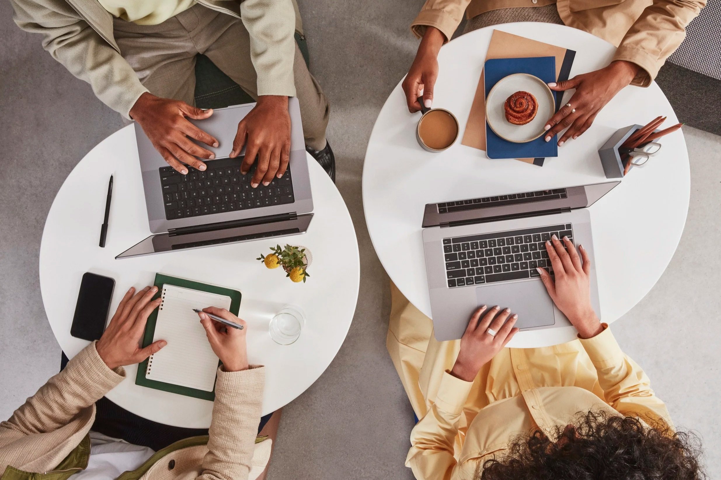 Overhead view of three people working at round white tables with laptops, notebooks, pens, and coffee mugs.