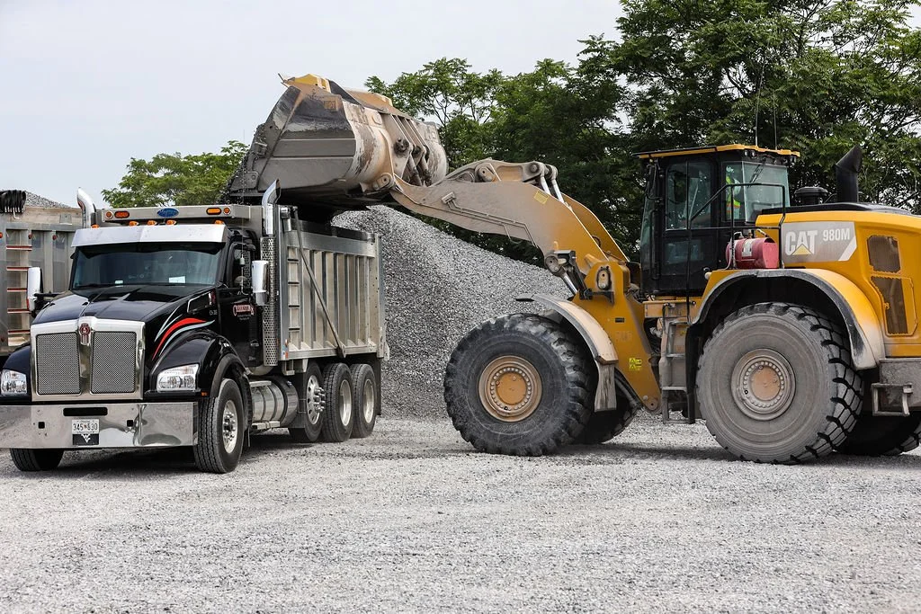 A large yellow Caterpillar front loader dumping gravel into a black and silver semi-truck on a gravel lot.