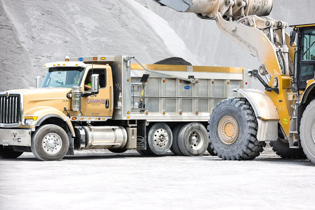 Creekstone yellow and silver mining dump truck being loaded with black material by a yellow front loader at a quarry site, highlighting heavy equipment operation, aggregate handling, and large-scale hauling services.