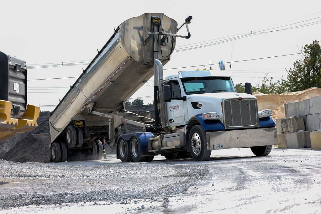 reekstone white semi-truck with a chrome grille and blue accents dumping gravel at a construction site, with surrounding gravel piles and concrete blocks, highlighting material delivery and heavy hauling services.