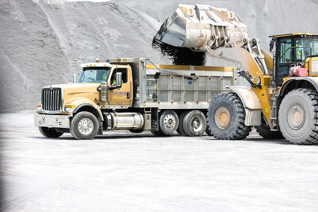 Creekstone dump truck unloading black material into a front loader at a quarry site with white dusty ground and gray rocky walls, highlighting heavy hauling and aggregate handling operations.