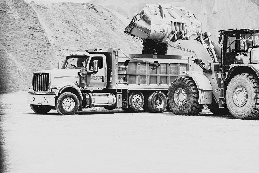 A large industrial dump truck unloading material into a cement mixer truck in a quarry or construction site.