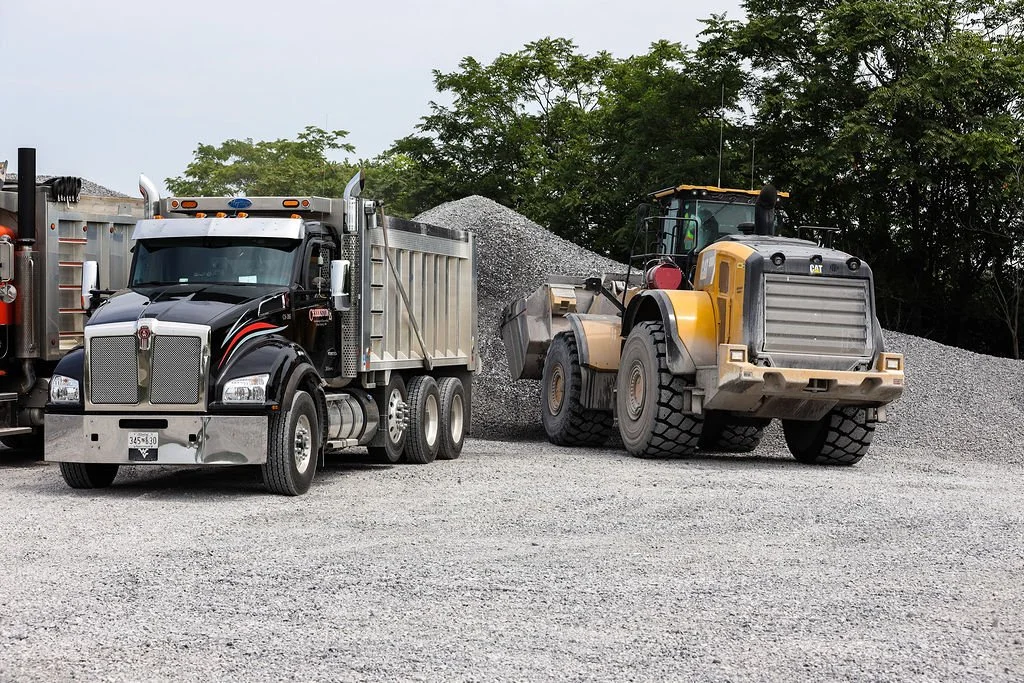 A Creekstone black semi-truck with silver and red accents and a gray dump truck working together at a construction site with gravel and green trees in the background.
