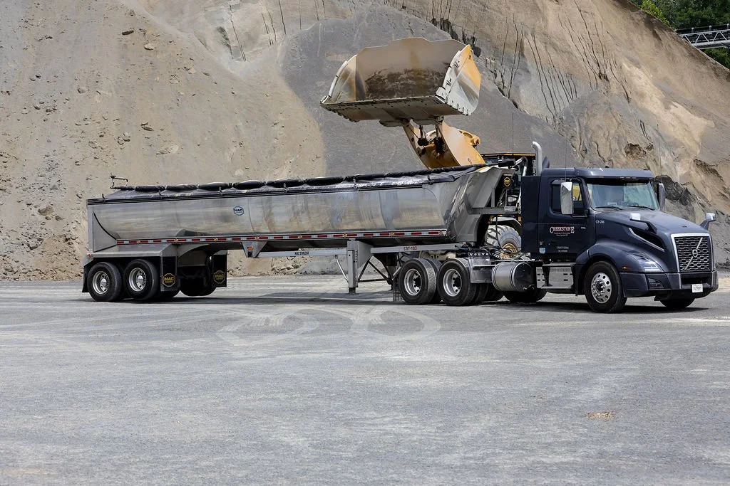 A large black semi-truck with a shiny silver trailer is in the process of unloading or loading gravel or sand into a mining or construction site. The truck's dump bed is tilted forward with gravel pouring out.