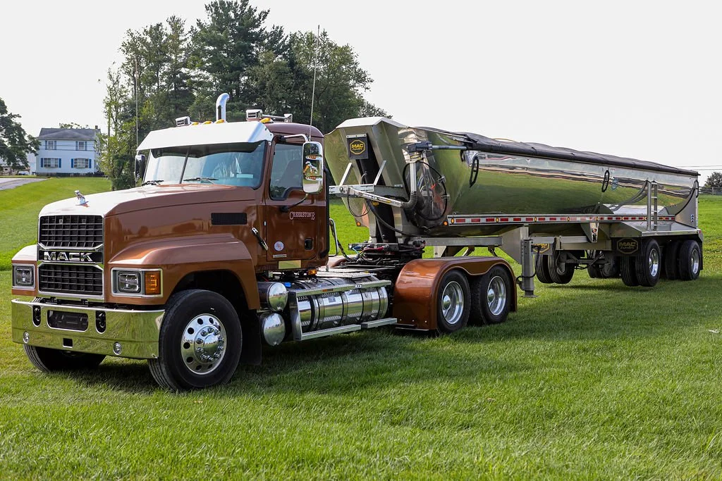 Creekstone brown Mack truck pulling a shiny metallic trailer parked on a grassy field with trees and a house in the background, highlighting heavy-duty trucking and hauling services.