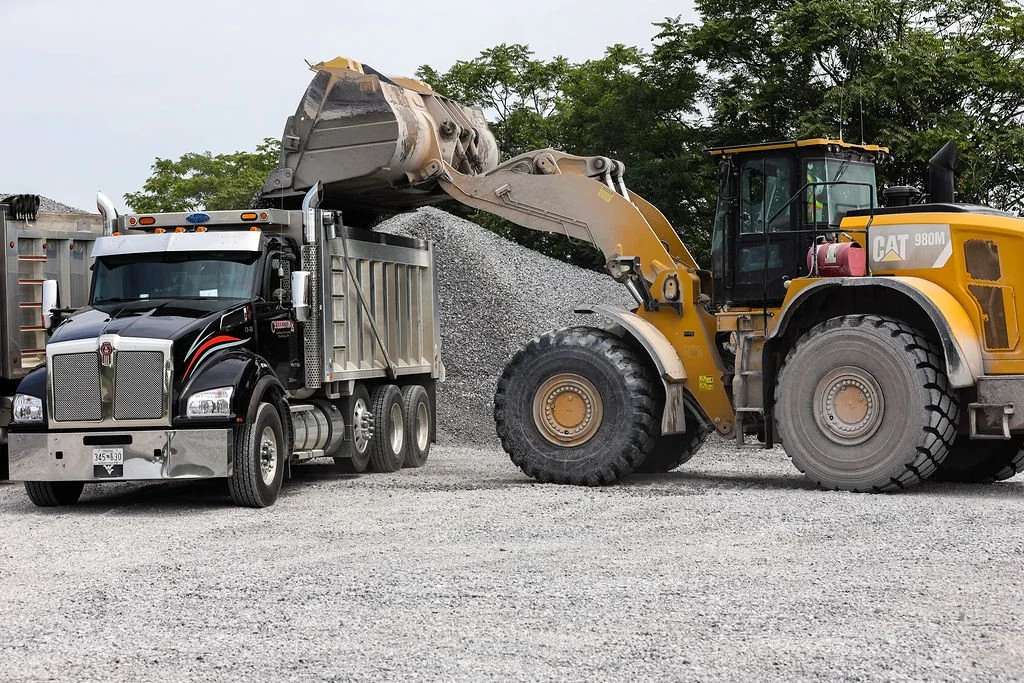 Creekstone yellow Caterpillar front loader dumping gravel into a black semi-truck with a silver container at an active construction site, showcasing heavy equipment operation and material hauling services.