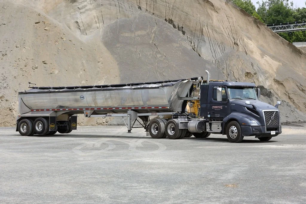 Creekstone black semi-truck with a silver tanker trailer parked on a gravel lot with a dirt or sand hill in the background, highlighting heavy-duty trucking and material transport services.