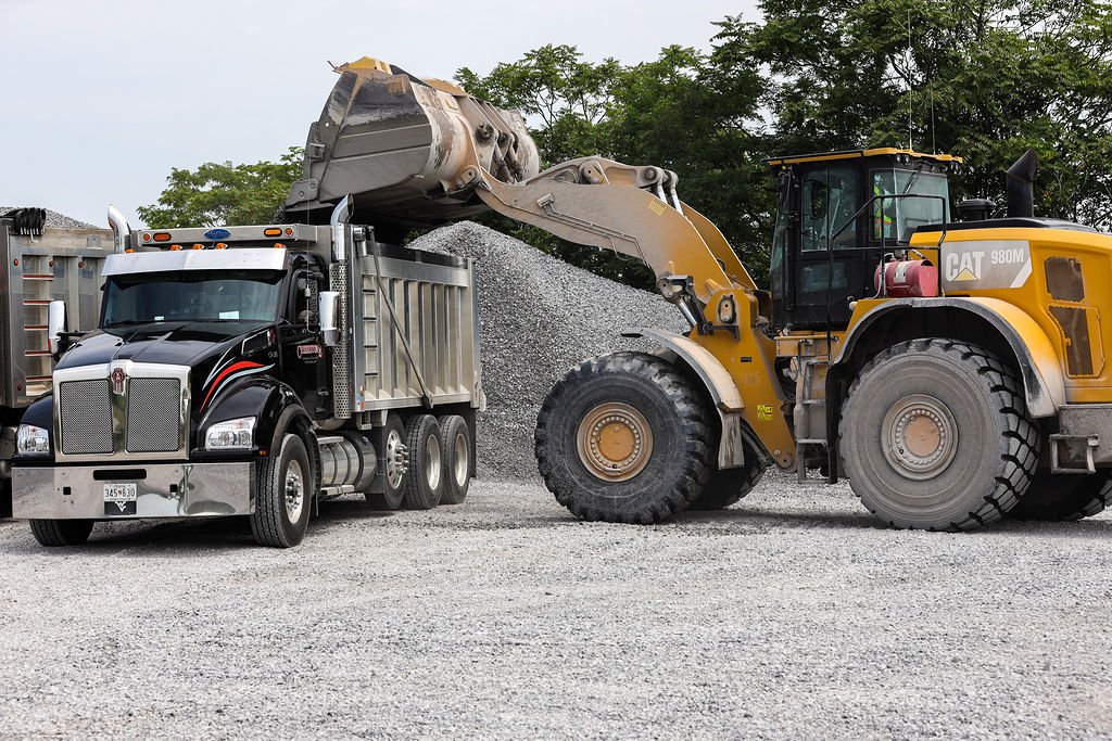 Creekstone front loader excavator dumping gravel into a black and silver dump truck at a construction site with trees in the background, highlighting heavy equipment operation and material hauling services.
