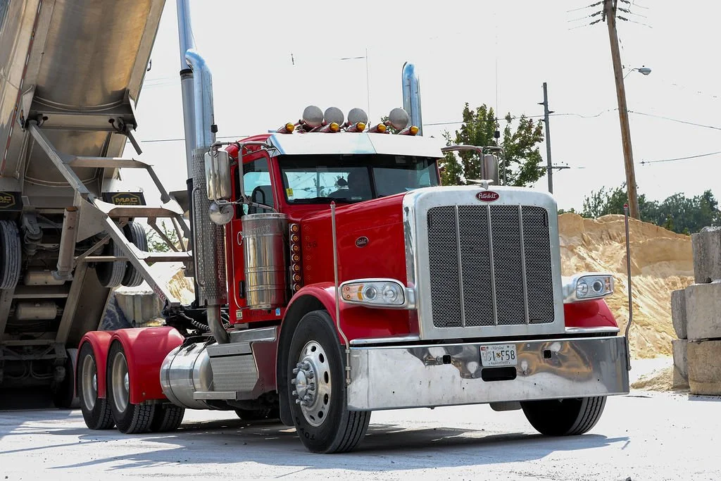 Creekstone red Peterbilt semi-truck with a large front grill parked beside a cement mixer truck at a construction site with dirt piles and utility poles in the background, highlighting heavy hauling and construction site operations.