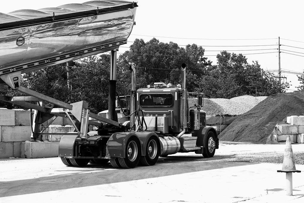 Creekstone semi-truck with a flatbed trailer parked at a construction site in a black-and-white image, with gravel and sand piles in the background and an orange safety cone in the foreground, highlighting trucking and construction site operations.