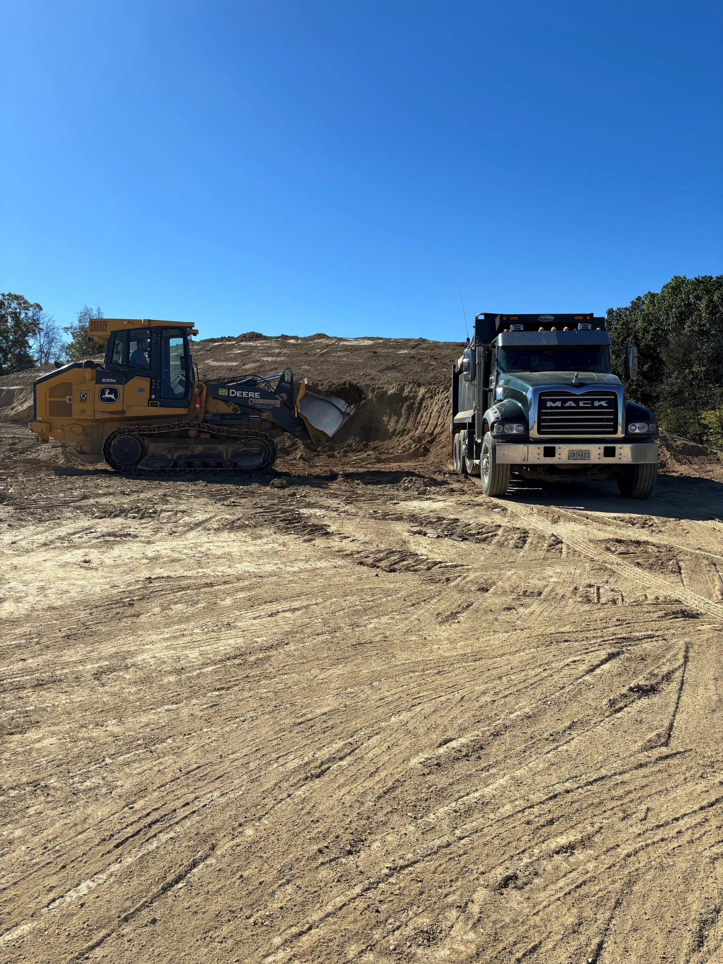 Creekstone construction site with a yellow bulldozer and a black Mack truck on uneven dirt ground under a clear blue sky, highlighting excavation, grading, and heavy hauling operations.