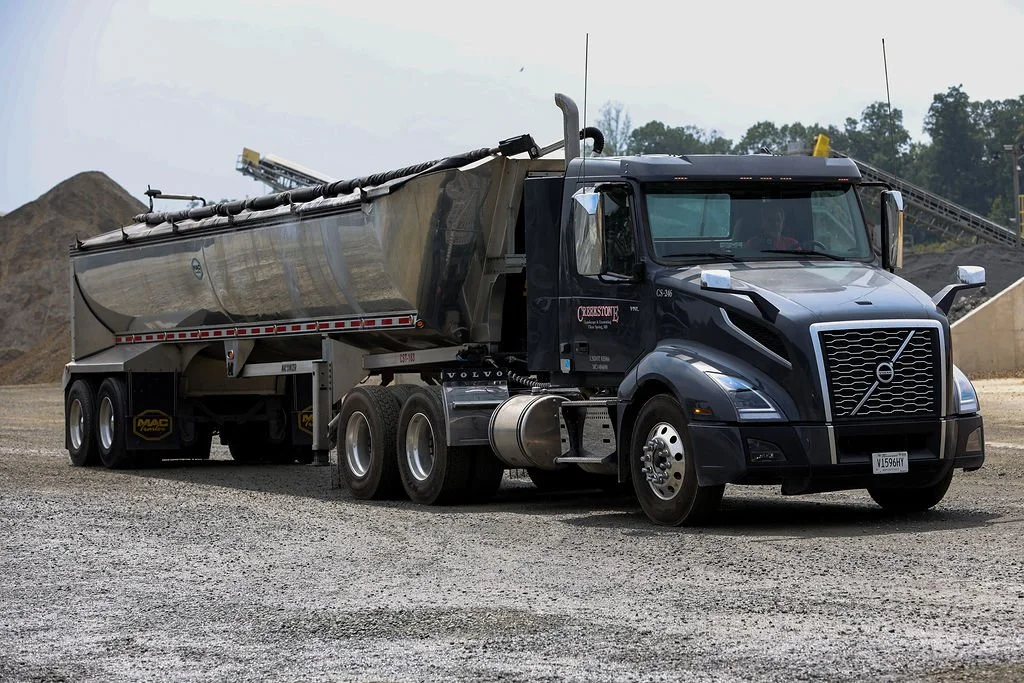 Creekstone black Volvo semi-truck with a reflective silver trailer parked on a gravel surface at a quarry-style construction site with gravel piles and trees in the background, highlighting heavy hauling and trucking services.