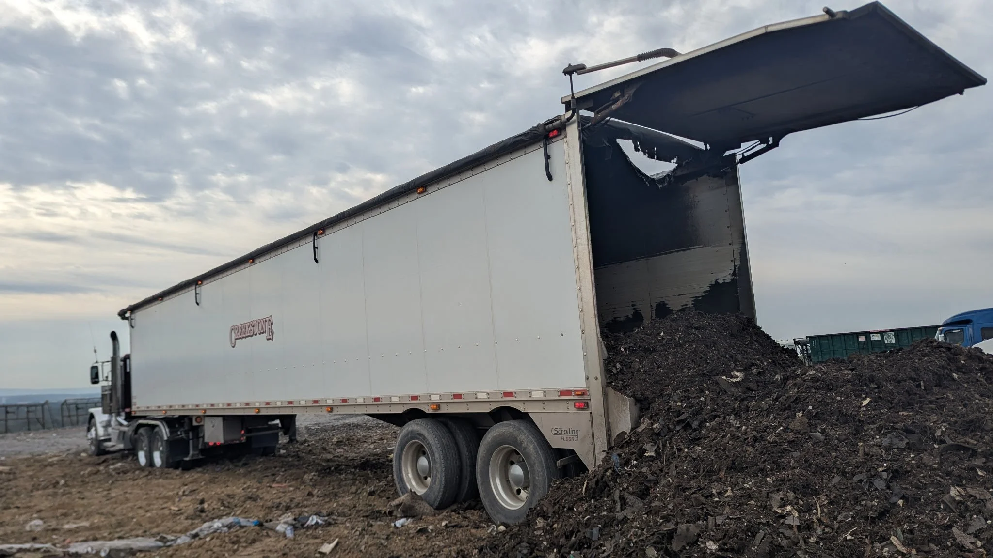 Creekstone white dump truck unloading a pile of dark soil or mulch at a construction or landscaping site under partly cloudy skies, highlighting heavy hauling and material delivery services.