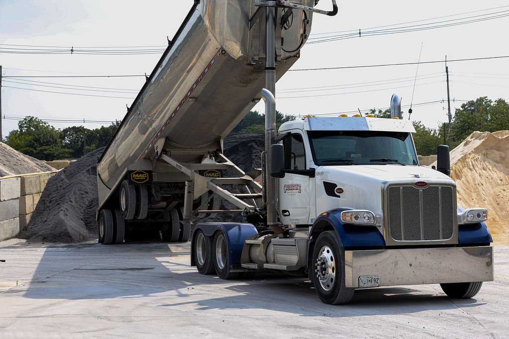 Creekstone white semi-truck with blue trim unloading black gravel from its tilted trailer at an active construction site, highlighting heavy hauling and material delivery services.