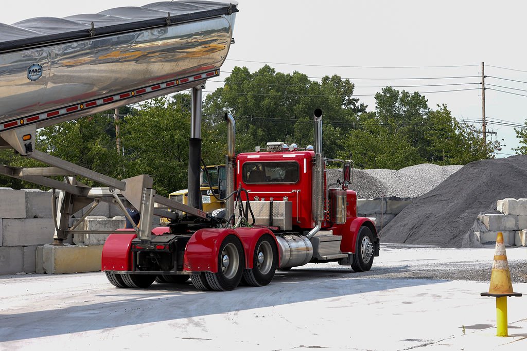 A red semi-truck with a large tanker trailer parked at a construction site, with concrete blocks and piles of gravel in the background.