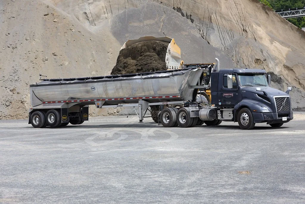 Creekstone black semi-truck with a silver dump trailer being loaded with dirt or gravel at a construction site with a dirt hill in the background, highlighting heavy hauling and material transport services.