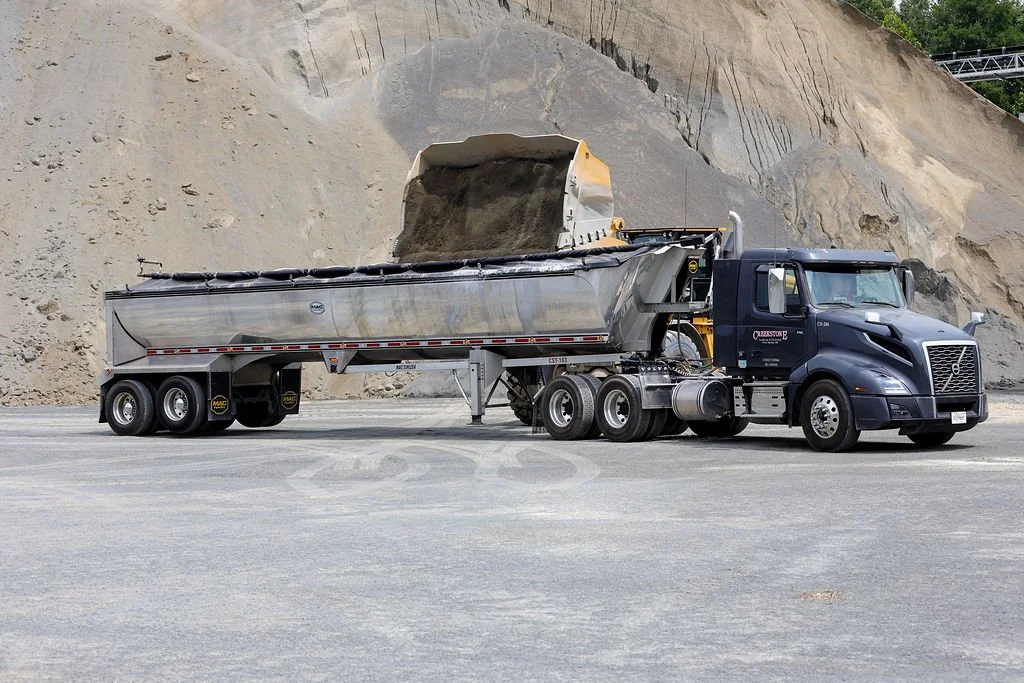 A Creekstone black Volvo semi-truck with a silver dump trailer transferring a load of dirt or sand on a construction site with a dirt hill in the background.