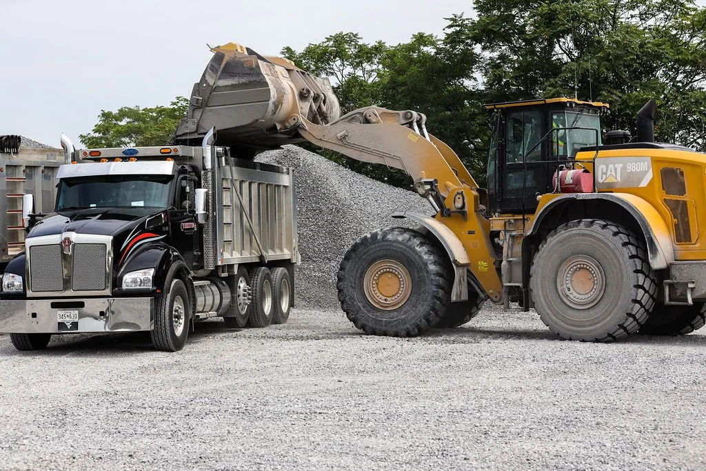 Creekstone yellow Caterpillar front loader transferring gravel into the bed of a black semi-truck on a gravel lot with trees in the background, showcasing heavy equipment operation and material hauling services.