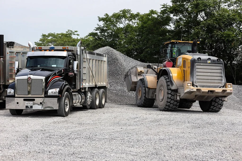 Creekstone black and silver dump truck filled with gravel parked beside a yellow bulldozer with a gravel pile in front of it on a gravel lot with trees in the background, highlighting heavy hauling and construction site operations.